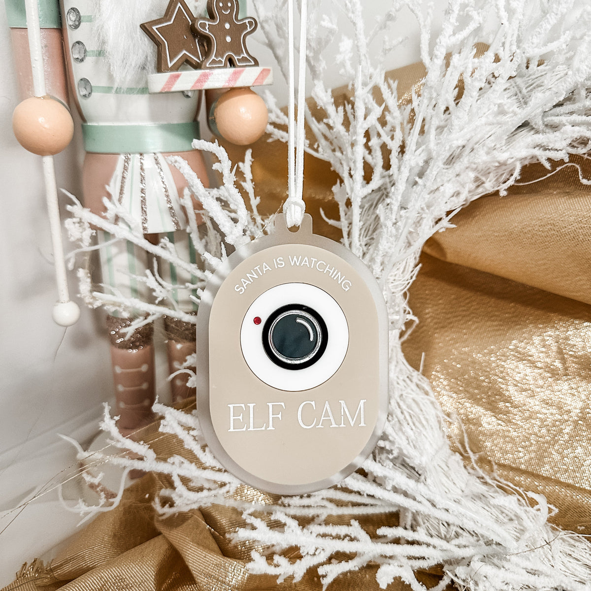 A round beige Christmas ornament with a camera graphic and the words 'ELF CAM' and 'SANTA IS WATCHING', hanging among white frosted branches and gold fabric, with a wooden nutcracker partially visible behind it.